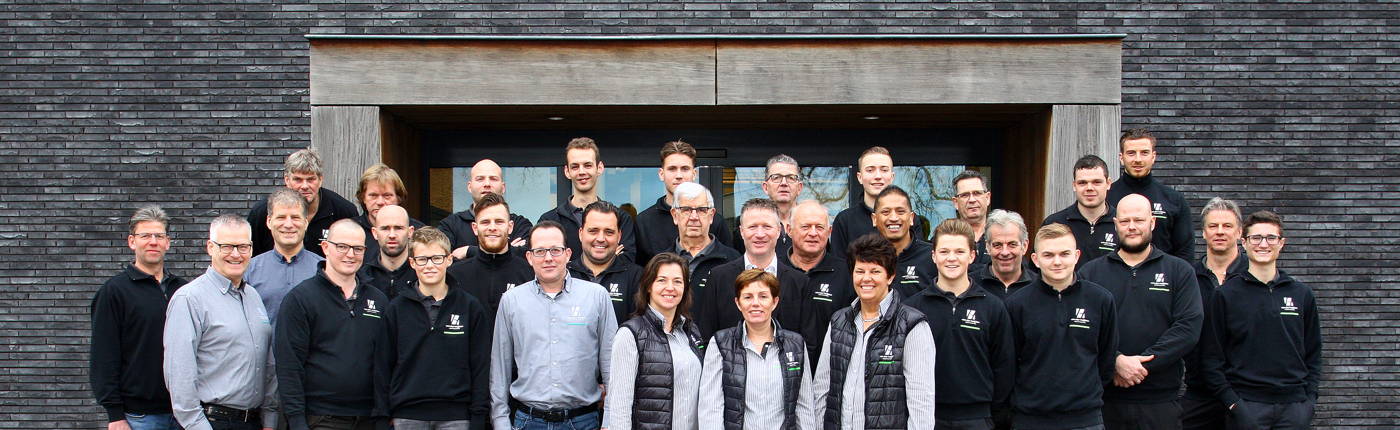 Groepsfoto van het team van Houthandel Van der Heijden, staand voor een moderne gevel met donkere stenen. Het team bestaat uit mannen en vrouwen, gekleed in bedrijfskleding met het logo van de houthandel. De medewerkers poseren lachend en stralen een prof
